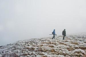 dos fotógrafos turísticos con trípode a mano caminando sobre una colina congelada con niebla. foto