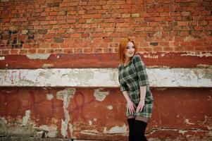 An outdoor portrait of a young pretty girl with red hair wearing checkered dress standing on the brick wall background in winter day. photo