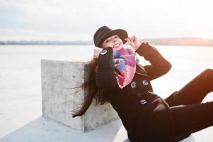 chica joven casual divertida en abrigo negro, bufanda y sombrero contra el río congelado en el clima soleado de invierno. foto