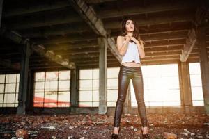 retrato de una joven morena linda con pantalones de cuero negro y blusa blanca posada en un lugar abandonado. foto