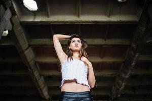 retrato de una joven morena linda con pantalones de cuero negro y blusa blanca posada en un lugar abandonado. foto