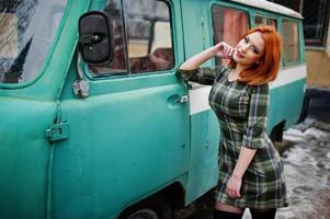 Young red haired girl posed on checkered dress background old retro cyan minivan.