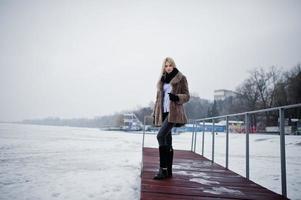 retrato de una joven rubia elegante con un abrigo de piel en el fondo del muelle río brumoso en el hielo invernal. foto