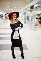 Fashion red haired girl wear on black dress and red hat with ladies backpack posed at trade shopping center. Photo toned style Instagram filters.