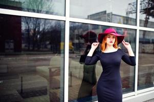Portrait of fashion red haired girl on red hat and black dress with bright make up posed against large window. Photo toned style Instagram filters.