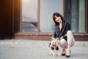 Brunette gypsy girl with yorkshire terrier dog posed against large windows house. Model wear on leather jacket and t-shirt with ornament, pants and shoes with high heels. photo