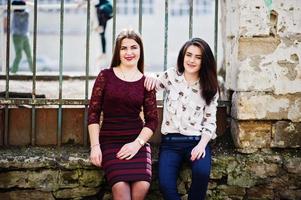 Two young teenages girl sitting against iron fence. Plump girl vs thin. Friends teen. photo