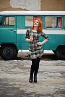 jovencita de pelo rojo con teléfono móvil y auriculares, vestida con un fondo de vestido a cuadros antiguo autobús minivan turquesa vintage. foto