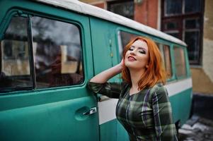 Young red haired girl posed on checkered dress background old retro cyan minivan. photo