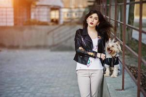 chica gitana morena con perro yorkshire terrier posó contra barandillas de acero. desgaste modelo en chaqueta de cuero y camiseta con adorno, pantalones. foto