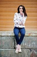 Young stylish brunette girl on shirt, pants and high heels shoes, sitting and posed background orange shutter. Street fashion model concept. photo