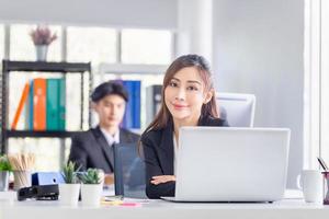 Attractive cheerful young business woman with laptop at office and blurred colleague sitting in the background