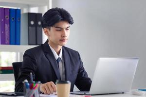 retrato de un joven oficinista sentado en su escritorio en la oficina, hombre de negocios con traje trabajando con una laptop en la oficina foto