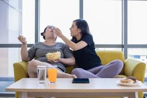 mujer con sobrepeso y niña asiática disfrutan comiendo comida en el sofá en casa