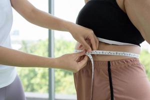 Young female trainer measuring fat layer of overweight woman with waistline at fitness photo