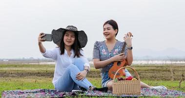 A group of Asian friends playing Ukelele and spending time making a picnic in the summer holidays.They are happy and selfie ,Relax time on holiday concept travel photo