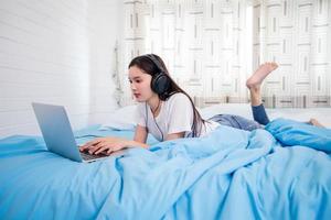 Asian happy young woman wear headphones and listening music song and watching website for shopping online from laptop  on bed at home photo