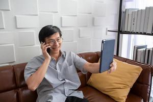 Senior Asian man playing tablet and video call on sofa in living room at home  Portrait of Asian elderly man is Relaxing and Happiness With playing tablet photo