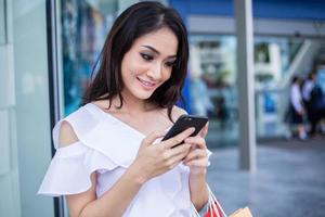 Beautiful asian young woman with shopping bags using her smart phone with smile while standing at the clothing store. Happiness, consumerism, sale and people concept photo