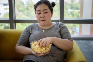 mujer con sobrepeso y niña asiática disfrutan comiendo comida y palomitas de maíz en el sofá en casa foto