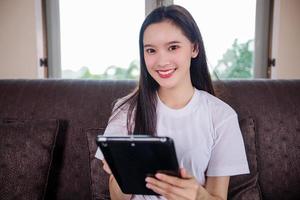 Asian young woman happy cheerful cute beautiful is playing digital pad tablet computer and sitting on the sofa at home, smiling lady using app reading electronic book relax in living room photo
