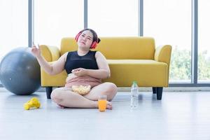 mujer asiática con sobrepeso usando tableta y. usando auriculares para escuchar música y ella está haciendo ejercicio en casa, una chica asiática disfruta comiendo comida y palomitas de maíz foto