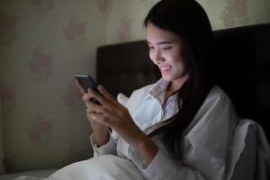 Asian woman using a mobile phone on night at the bed.She smiles and enjoys at home. photo