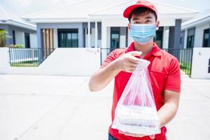 Asian delivery servicemen wearing a red uniform with a red cap and face mask handling food boxes in plastic bags to give to the customer in front of the house. Online shopping and Express delivery photo