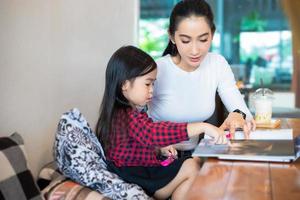 Asian mothers are teaching their daughters to read a book and use notebooks and technology for online learning during school holidays at home. Educational concepts and activities of the family photo