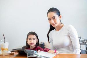 An Asian mother is teaching her daughter to read a book during the semester break on the living table and having cold milk on the table at home. Educational concepts and activities of the family photo