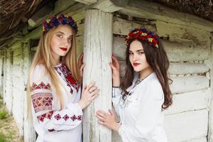dos jóvenes mujeres hermosas con el pelo largo en blusas ucranianas y en una corona en una aldea étnica al aire libre en kiev, ucrania foto