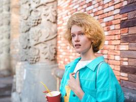 young cheerful curly redhead woman in blue jeans jacket and straw hat holding cup of coffee and surprised by landmarks. Fun, summer, travel, fashion, youth concept photo