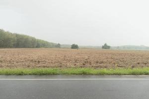 Horizontal view of wet asphalt road in Thailand. Environment of rainy time. Background of seeding of cassava plantation. and rubber trees far way. Under the dark sky. photo