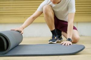 un deportista asiático activo desenrolla una colchoneta de ejercicio en el suelo, un cuerpo sano y en forma de un deportista asiático que prepara una colchoneta de yoga para el peso corporal y el ejercicio cardiovascular. foto