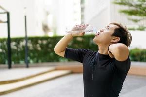 deportista asiático activo relajándose y bebiendo agua en una botella después de correr o hacer ejercicio al aire libre. un hombre asiático bebe agua durante el descanso del ejercicio. un hombre sano que se esfuerza por hacer ejercicio cardiovascular. foto