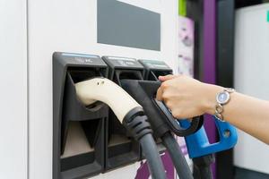 feliz mujer asiática sosteniendo un conector de carga dc - ccs tipo 2 ev en la estación de carga ev, mujer preparando un ev - conector de carga de vehículos eléctricos para recargar un vehículo. foto