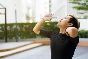 deportista asiático activo relajándose y bebiendo agua en una botella después de correr o hacer ejercicio al aire libre. un hombre asiático bebe agua durante el descanso del ejercicio. un hombre sano que se esfuerza por hacer ejercicio cardiovascular. foto