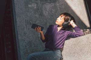 Happy young asian woman listening to music with headphones via smartphone and having fun while sitting side the street photo