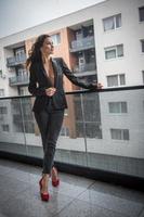 Hermosa mujer morena glamurosa con chaqueta negra posando en un balcón moderno con una vista increíble de la ciudad. Retrato de una elegante mujer de moda con piernas largas, jeans negros en el balcón foto