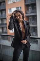 Hermosa mujer morena glamurosa con chaqueta negra posando en un balcón moderno con una vista increíble de la ciudad. Retrato de una elegante mujer de moda con piernas largas, jeans negros en el balcón foto