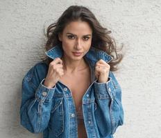 Hermosa mujer morena glamurosa con chaqueta azul posando en un balcón moderno con una vista increíble de la ciudad. Retrato de una elegante mujer de moda con cabello largo, jeans negros en el balcón foto