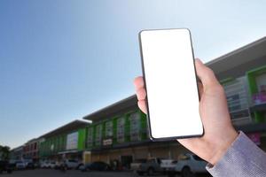 White blank touch screen smartphone holding in hand with blurr modern  buildings background. Close up view of right hand with mobile device. Mock up, copyspace, template and technology concept. photo