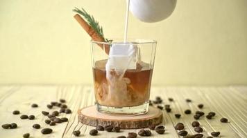 pouring milk in black coffee glass with ice cube, cinnamon and rosemary on wood background video