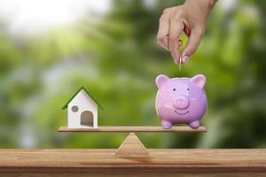 Model house and hand holding a coin in a pig piggy bank placed on the scales and blurred green background. Real estate ideas and future investment business plans.
