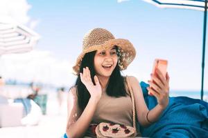viajar mujer asiática relajarse en el café de la playa y usar el teléfono móvil para videollamadas a través de Internet en línea. foto