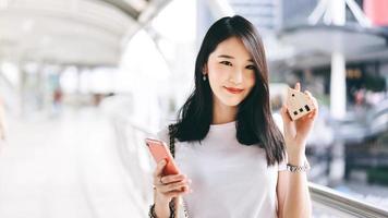 Mujer asiática que trabaja en una empresa adulta joven que sostiene un teléfono móvil y un modelo de casa para el concepto de solicitud de seguro en línea.
