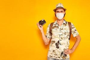 a male tourist is a traveler on vacation in medical mask with camera in his hand, backpack on his shoulders yellow isolated background with space for text. concept - people, adventures, sightseeing photo
