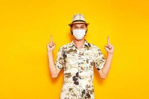 male tourist in medical mask points with his hands up at an empty advertising area above him. isolated yellow background. copy space. concept of people, travel, vacation, protection from covid19 virus