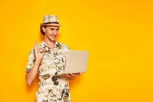 male tourist traveler on vacation with laptop in his hand shows OK sign, everything is fine via video link. isolated on yellow background with space for text. concept people, communication, recreation photo