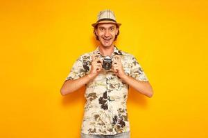 an excited male tourist traveler on vacation in hat and Hawaiian shirt holds camera in front of him and happy to see resulting frame. isolated on yellow background with space for text. concept people photo
